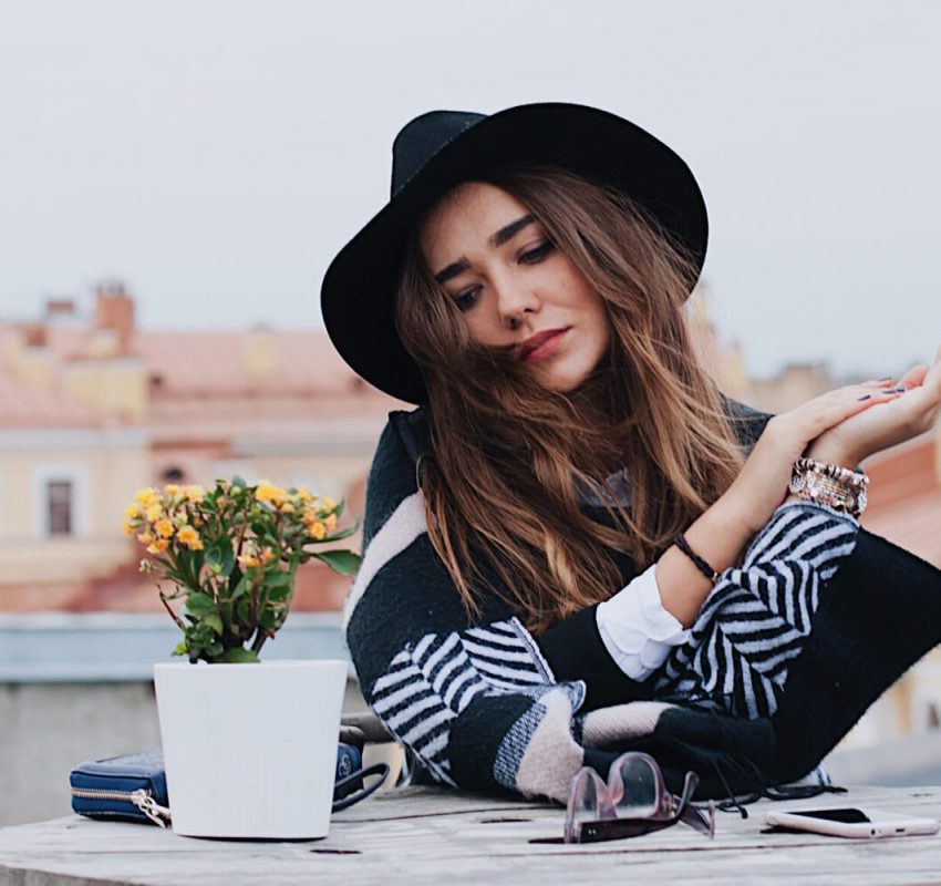 A woman wearing a black hat and striped shawl sits at an outdoor table with a potted plant, sunglasses, and a small bag. She looks down with her hand raised, city buildings blurred in the background.
