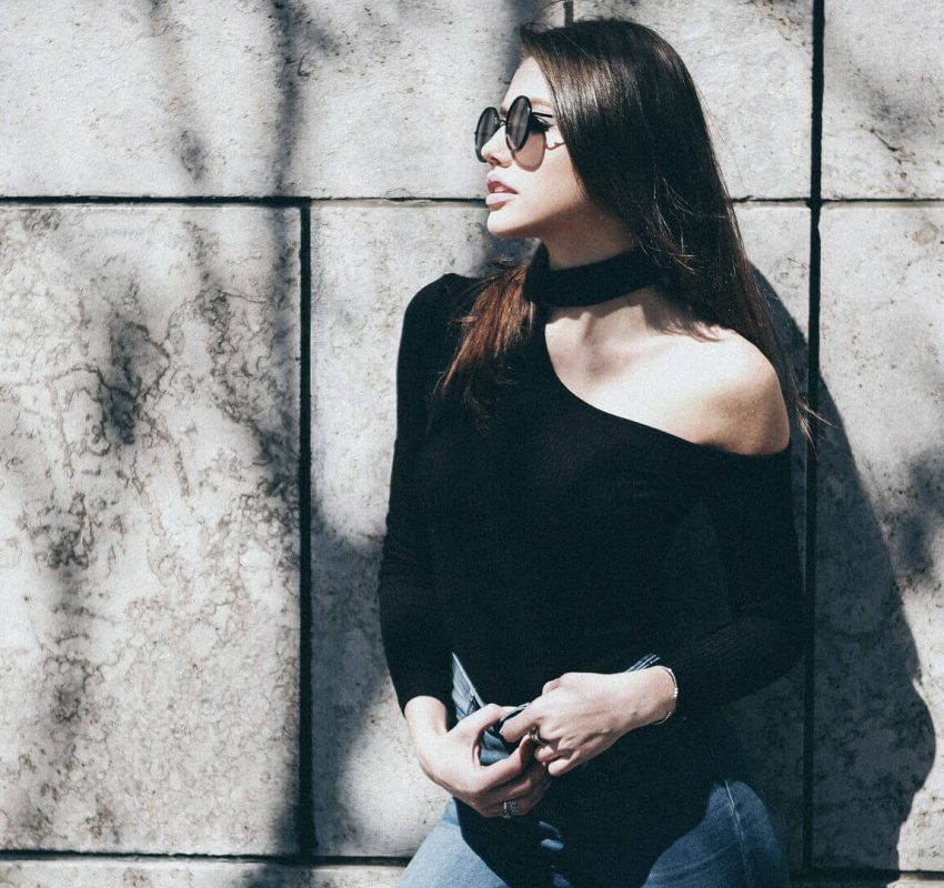A woman wearing sunglasses, a black off-the-shoulder top, and jeans stands against a sunlit stone wall with shadows of branches cast across it. She is looking to the side and holding a smartphone.