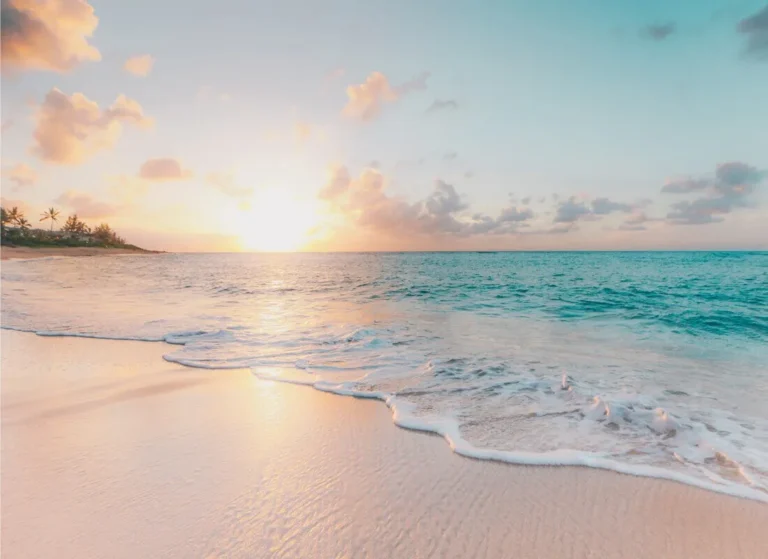 A serene beach at sunrise, with gentle waves washing onto soft sand. The sky is filled with pastel clouds, and the sun is just above the horizon, casting a warm glow over the ocean and shoreline.