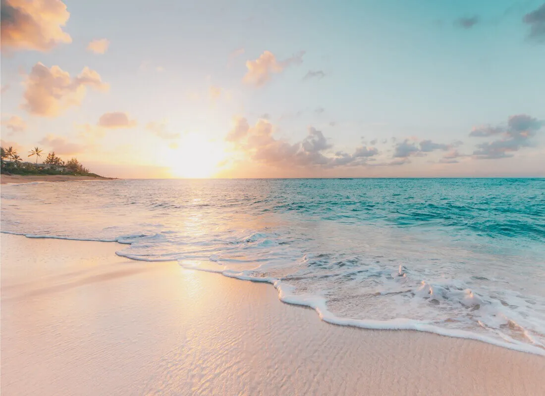 A serene beach at sunrise, with gentle waves washing onto soft sand. The sky is filled with pastel clouds, and the sun is just above the horizon, casting a warm glow over the ocean and shoreline.