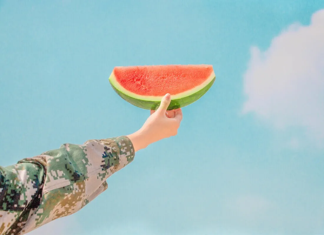 A person wearing a camouflage sleeve holds up a slice of watermelon against a bright blue sky with a few wispy clouds.