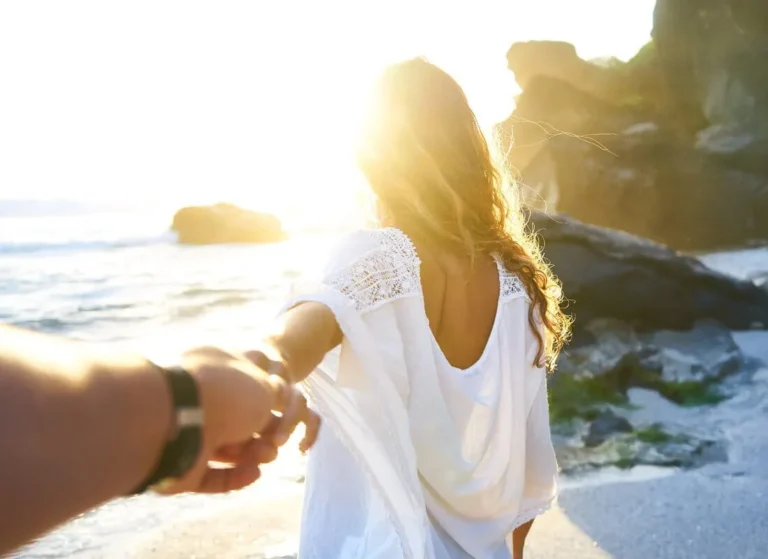 A woman with long hair in a white dress stands on a sunny beach, holding someone’s hand and leading them forward toward the shoreline, with rocks and sunlight in the background.