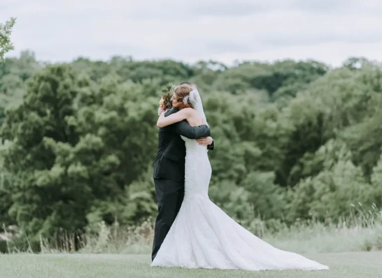 A bride in a white gown and veil hugs a groom in a black suit on a grassy field, with lush green trees in the background under a cloudy sky.