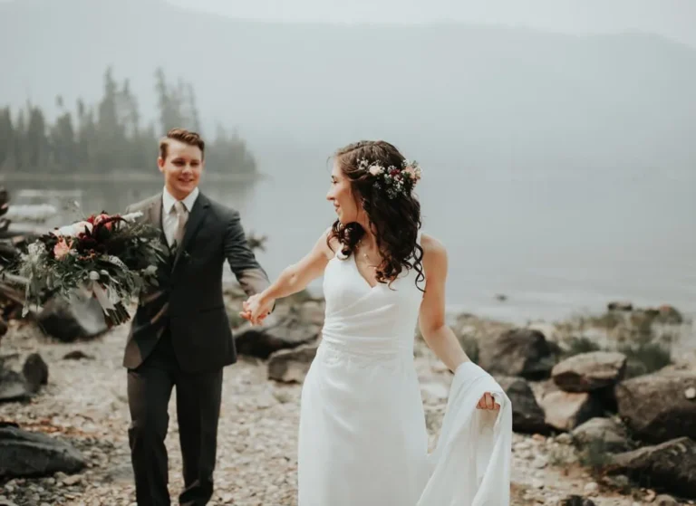 A bride in a white dress holding her skirt smiles and leads a groom in a dark suit holding a bouquet along a rocky lakeshore with misty mountains in the background.