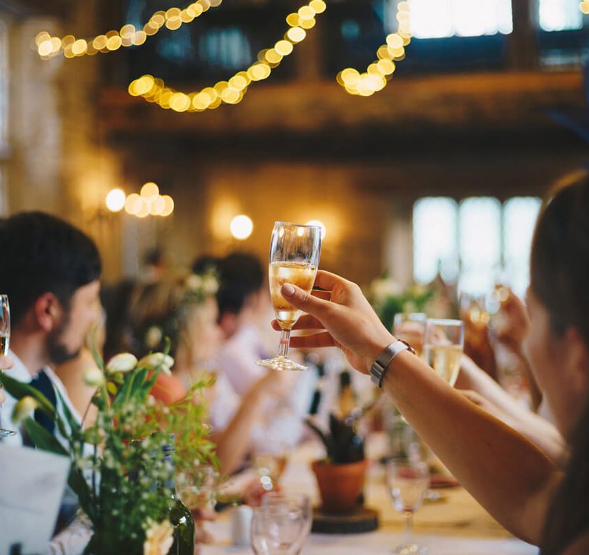People seated around a table raise glasses in a toast at a warmly lit indoor gathering, with blurred string lights and floral centerpieces adding to the festive atmosphere.