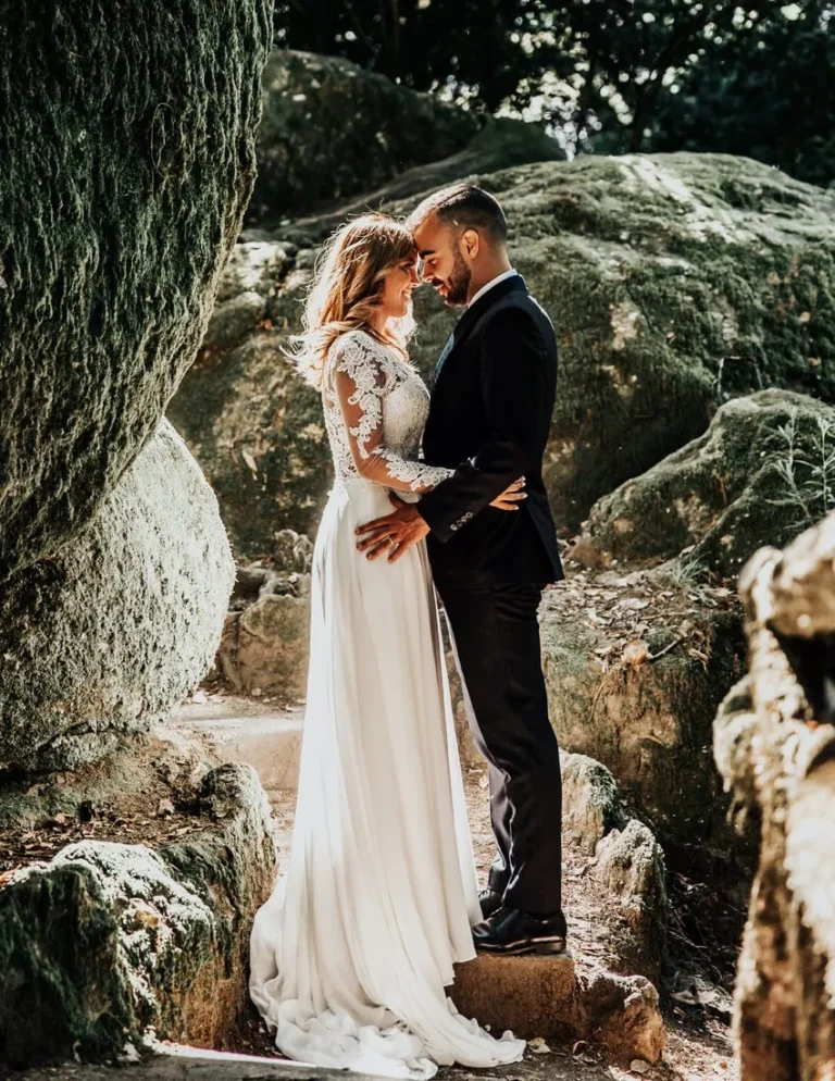 A bride and groom stand close together on a rocky path outdoors, embracing and smiling softly, surrounded by large boulders and dappled sunlight. The bride wears a long white dress; the groom wears a black suit.
