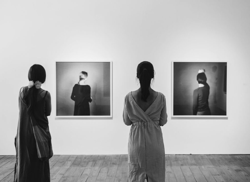 Two people stand in an art gallery, facing two framed black-and-white photographs on a white wall. The images depict individuals facing away from the camera. The scene is in black and white.