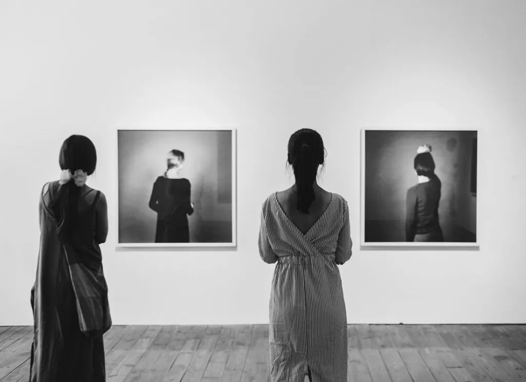 Two people stand in an art gallery, facing two framed black-and-white photographs on a white wall. The images depict individuals facing away from the camera. The scene is in black and white.