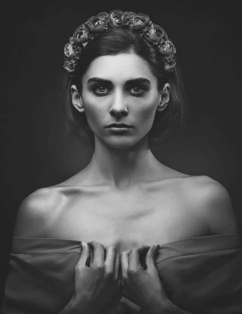 A black and white portrait of a woman with short hair and a floral headpiece. She looks directly at the camera, holding a draped fabric close to her chest, with bare shoulders and a serious expression.