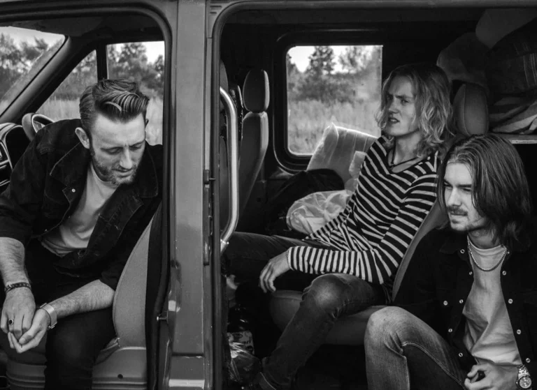 Three young adults sit inside a van, looking relaxed and thoughtful. One sits in the open doorway, another in the back seat, and the third reclines inside. The photo is black and white, giving it a moody, candid feel.