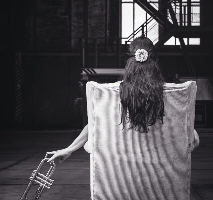 A woman with long hair and a flower hair accessory sits in an armchair, facing away. She holds a trumpet in one hand, resting it beside the chair. Industrial stairs and large windows are visible in the background.