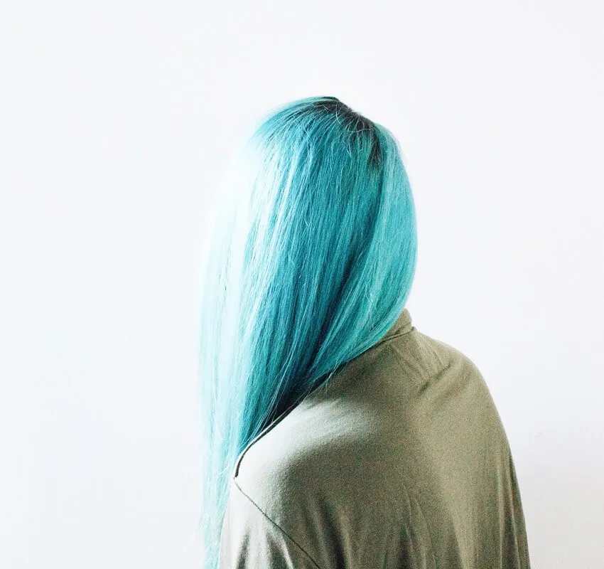A person with long, straight turquoise hair is seen from behind, wearing a loose, light grayish-green top, against a plain white background.