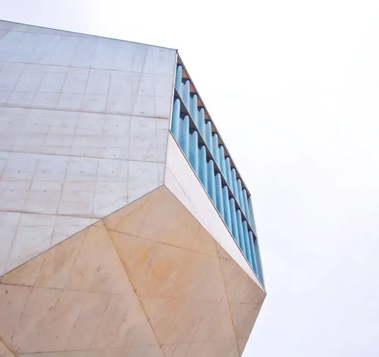A modern building with geometric, angular shapes and a concrete facade, featuring large vertical blue-tinted windows on the upper section against a pale, overcast sky.