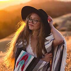 A young woman with long hair and glasses, wearing a black hat and a patterned blanket shawl, stands outdoors in a sunlit field at sunset.