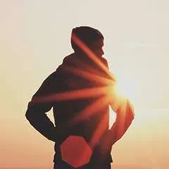 Silhouette of a person standing outdoors with hands on hips, facing the bright setting sun, which creates a warm glow and sun rays in the background.