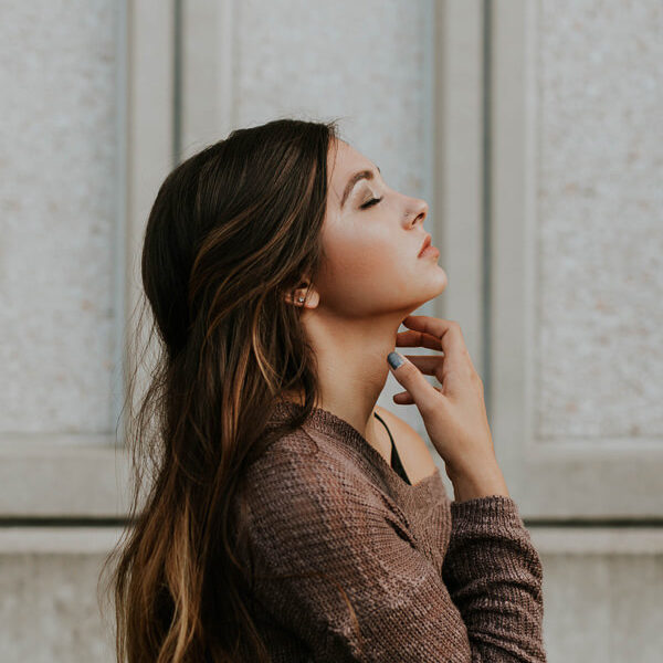 Woman with long brown hair and closed eyes poses in profile, gently touching her neck with her hand, wearing a textured brown sweater, standing against a light-colored wall with vertical panels.