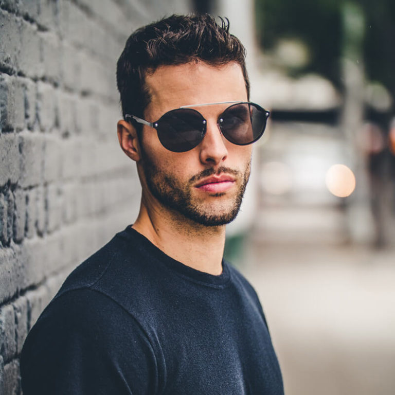 A man with short dark hair and a trimmed beard wears dark sunglasses and a black shirt, standing against a gray brick wall on a city street. The background is blurred.