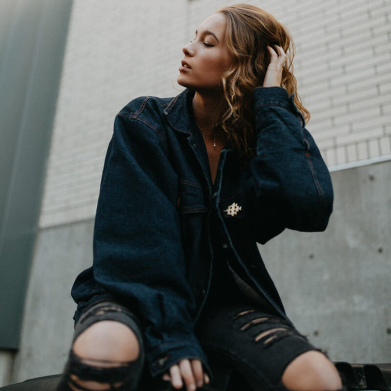 A woman with wavy hair sits outdoors against a brick wall, wearing a loose dark denim jacket and ripped black jeans, looking to the side with one hand in her hair.