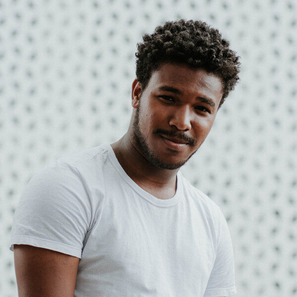 A young man with short curly hair and a trimmed beard, wearing a plain white t-shirt, stands in front of a light-colored, patterned background, looking slightly towards the camera with a subtle smile.