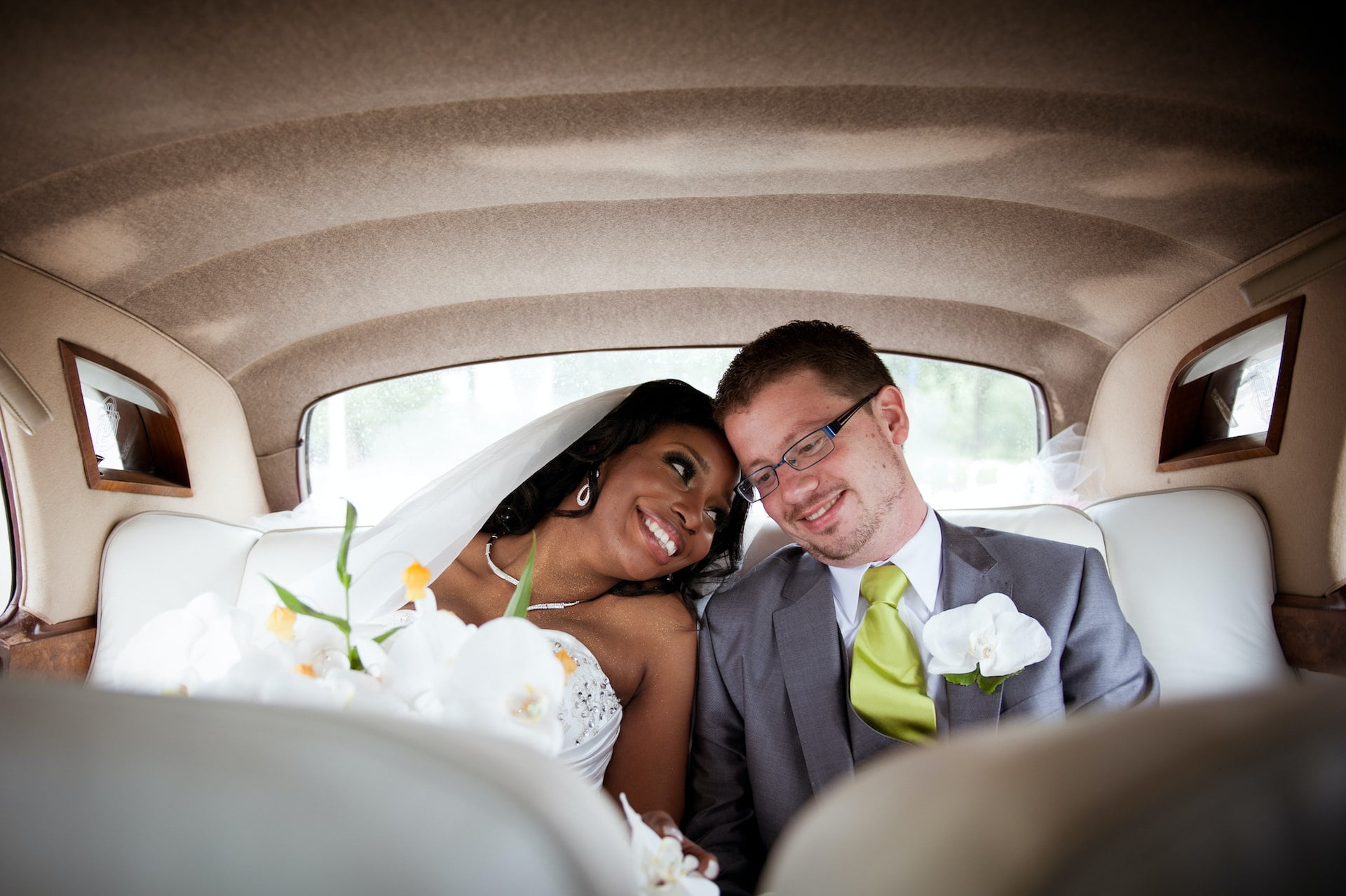 A smiling bride and groom are sitting closely together in the back of a vintage car. The bride wears a white dress and veil, holding a bouquet of white flowers. The groom is in a gray suit with a lime green tie, and they are leaning against each other.