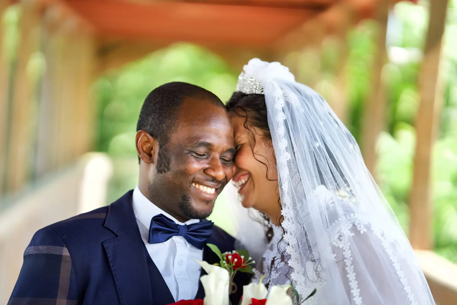 A joyful couple on their wedding day, the groom in a navy suit and bow tie, and the bride in a white dress with a veil and tiara. They are smiling and embracing, holding a bouquet of red and white flowers, against a blurred outdoor background.