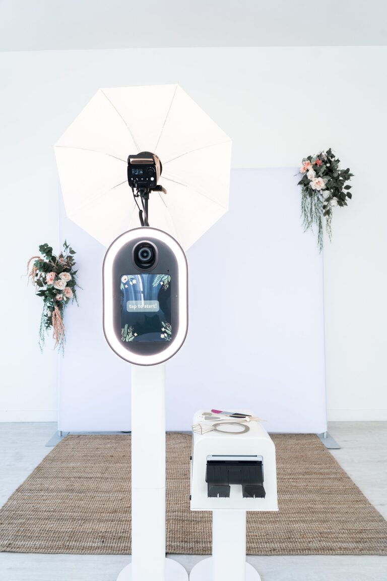 A photo booth setup with a camera mounted under a bright umbrella light. Floral decorations adorn the backdrop. A white machine with a slot, likely for printing photos, stands in front on a woven rug. The background is minimalist and bright.