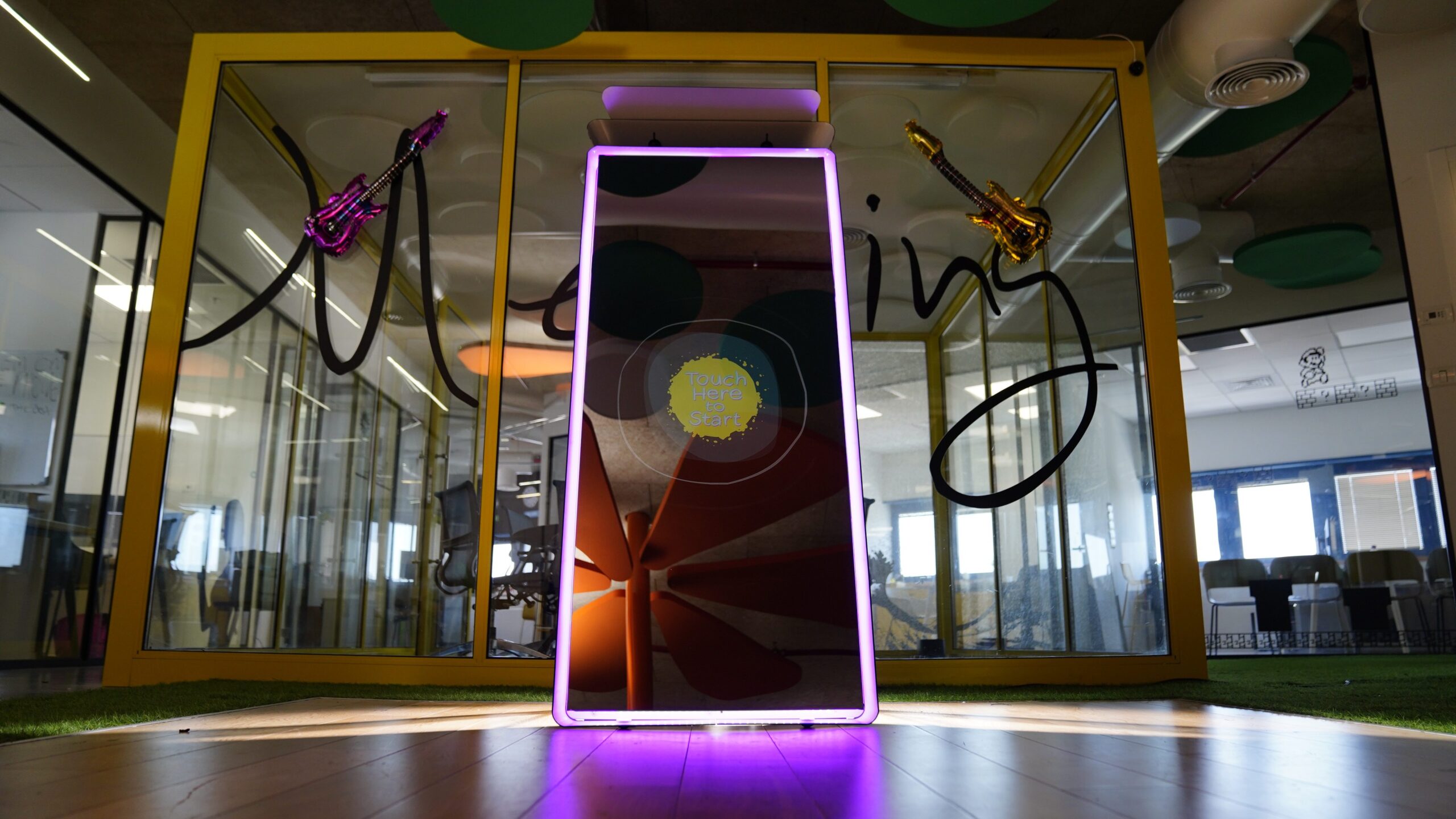 A tall interactive mirror with a neon purple frame stands in a brightly lit room. Behind it, glass walls feature black script and hanging gold balloon decorations shaped like guitars. The floor is wooden, and office space is visible in the background.