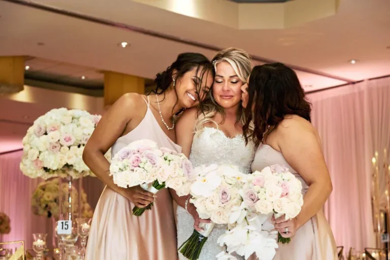 Three women in elegant dresses, including a bride in white, stand close together, smiling and embracing while holding large bouquets of white and pale pink flowers at a decorated indoor wedding venue.