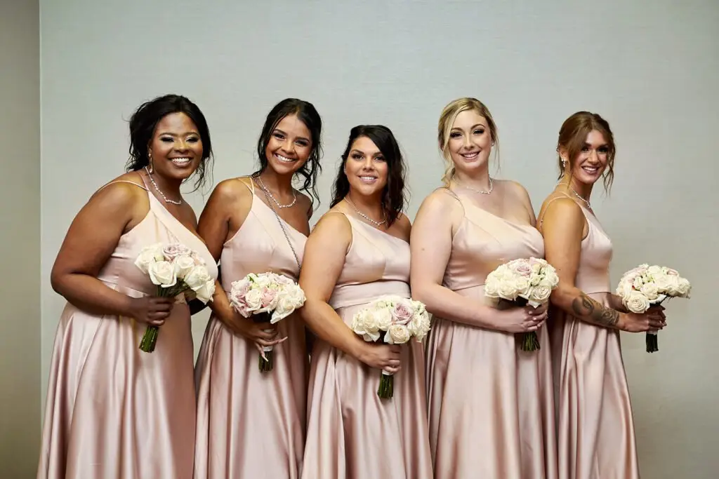 Five bridesmaids in matching pale pink dresses stand side by side, smiling and holding bouquets of white and blush flowers against a plain light background.