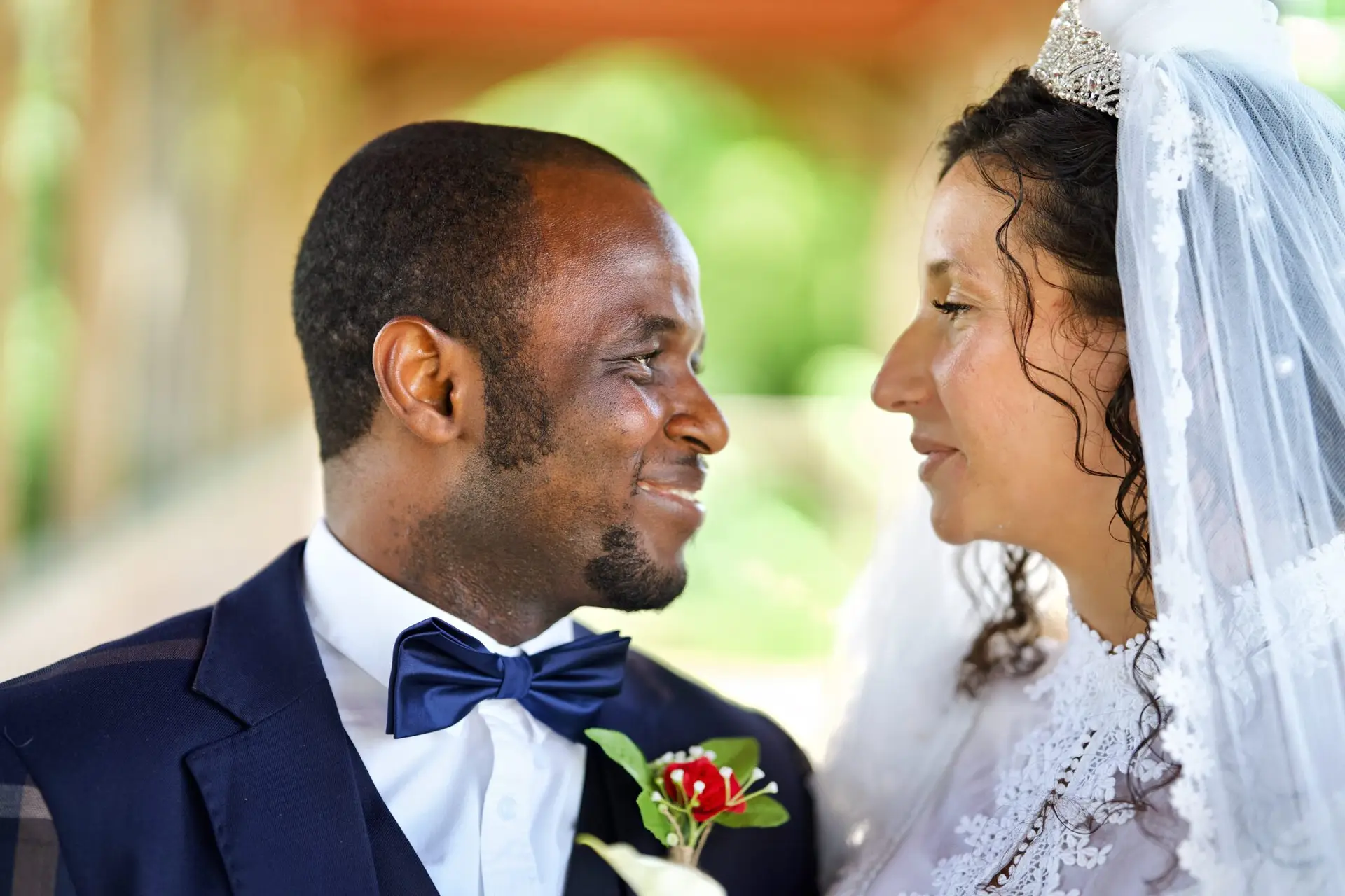 A bride and groom stand close together, facing each other and smiling. The groom wears a dark suit with a bow tie and boutonniere, while the bride wears a white dress, veil, and tiara. The background is softly blurred.