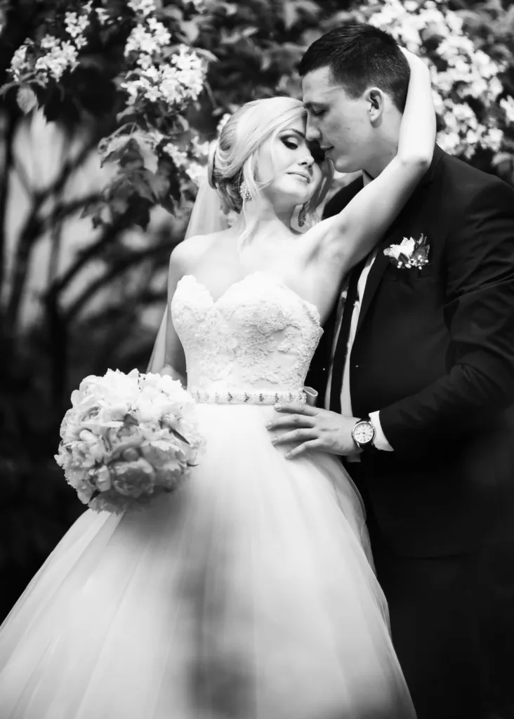 A bride and groom embrace lovingly outdoors, the bride holding a bouquet and wearing a strapless gown while the groom wears a suit. They share an intimate moment beneath blooming branches. Black and white photo.
