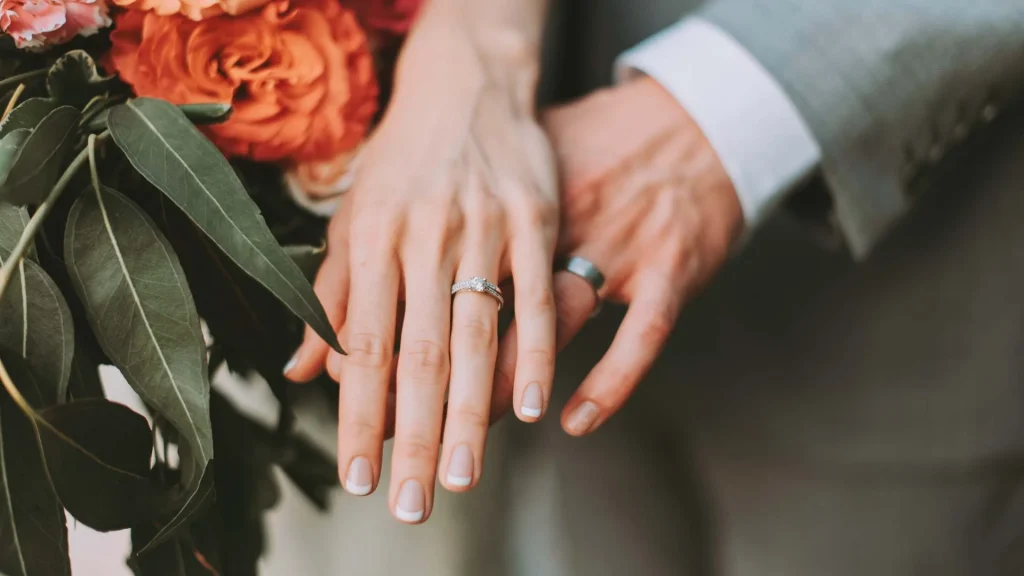 Close-up of two hands with wedding rings, one with a diamond engagement ring, gently touching over a bouquet of orange and pink flowers with green leaves. Both hands have light skin tones.
