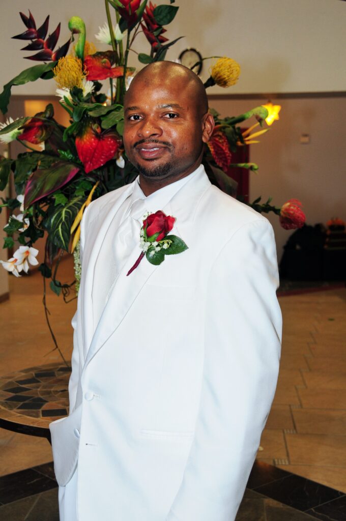 A man in a white suit with a red rose boutonnière smiles and poses indoors in front of a large, colorful floral arrangement.