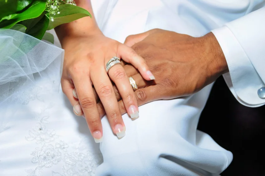 Close-up of two hands, one light-skinned and one dark-skinned, gently resting on each other. Both wear wedding rings, and part of a white wedding dress and bouquet are visible in the background.
