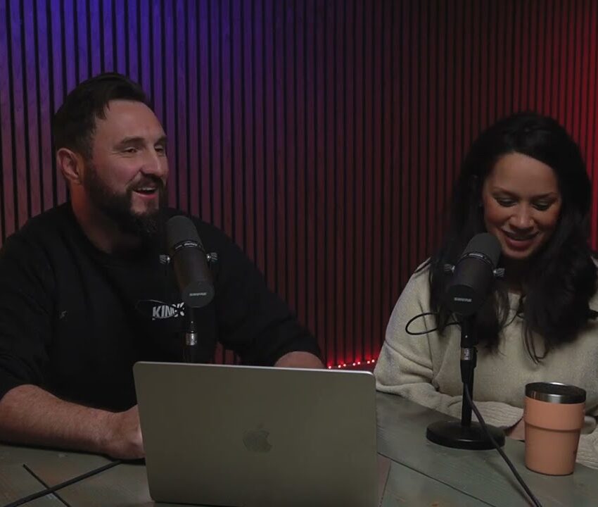 A man and a woman sit at a table with microphones, recording a podcast. The man smiles while the woman looks down and smiles. A laptop and a mug are on the table, with a wooden wall and shelves in the background.