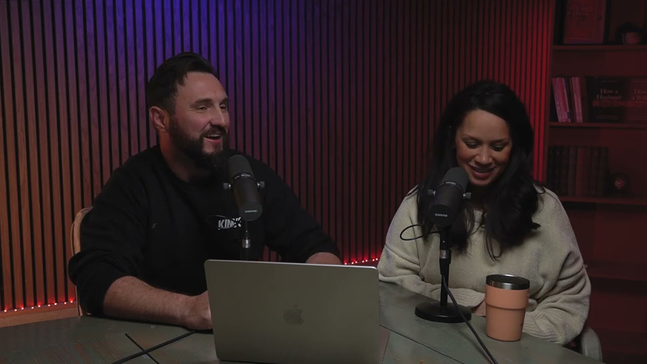 A man and a woman sit at a table with microphones, recording a podcast. The man smiles while the woman looks down and smiles. A laptop and a mug are on the table, with a wooden wall and shelves in the background.