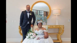 A bride in a white gown and tiara sits holding a bouquet, while a groom in a black tuxedo stands beside her, both smiling. They are in front of a gold table and round mirror, with a lamp and decorative items behind them.