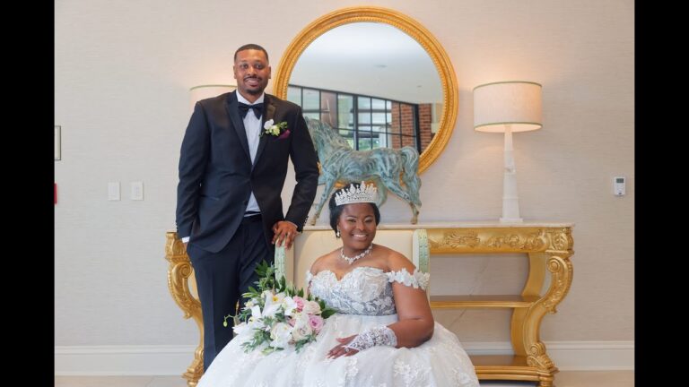 A bride in a white gown and tiara sits holding a bouquet, while a groom in a black tuxedo stands beside her, both smiling. They are in front of a gold table and round mirror, with a lamp and decorative items behind them.