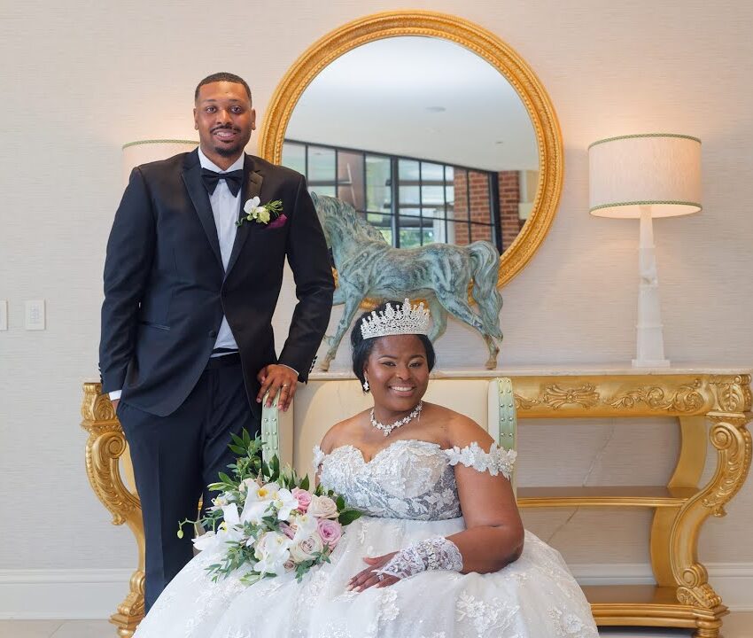 A bride in a white gown and tiara sits holding a bouquet, while a groom in a black tuxedo stands beside her, both smiling. They are in front of a gold table and round mirror, with a lamp and decorative items behind them.