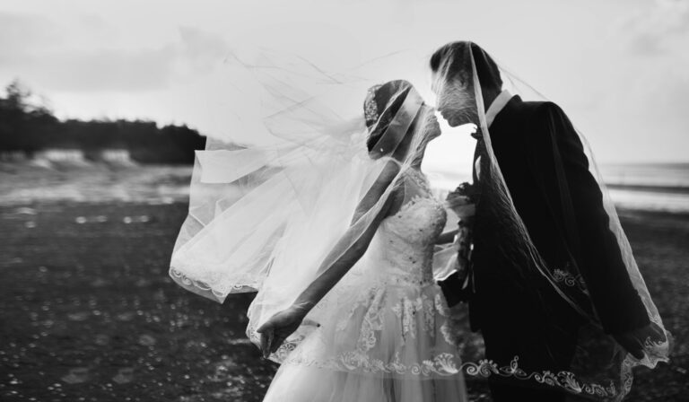 A bride and groom stand on a beach, facing each other closely under the bride’s flowing veil, sharing an intimate moment. The image is black and white, with soft light and a romantic atmosphere.