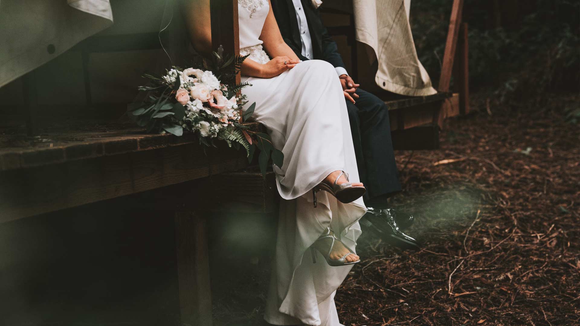 A bride in a white dress and a groom in a suit sit closely on a wooden bench outdoors. The bride holds a bouquet of flowers with pink and white blooms. The ground is covered in brown pine needles, and greenery surrounds them.